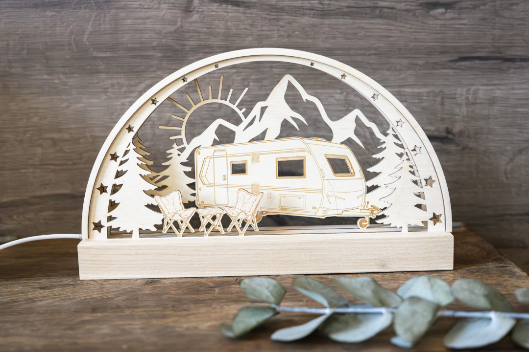 LED-Schwibbogen aus Holz mit Wohnwagen-Motiv vor Bergkulisse mit Sonne, Sternen und Tannen, davor Campingstühle und Tisch, warmweiß beleuchtet, mit Schriftzug „Camperleben" auf der Holzleiste